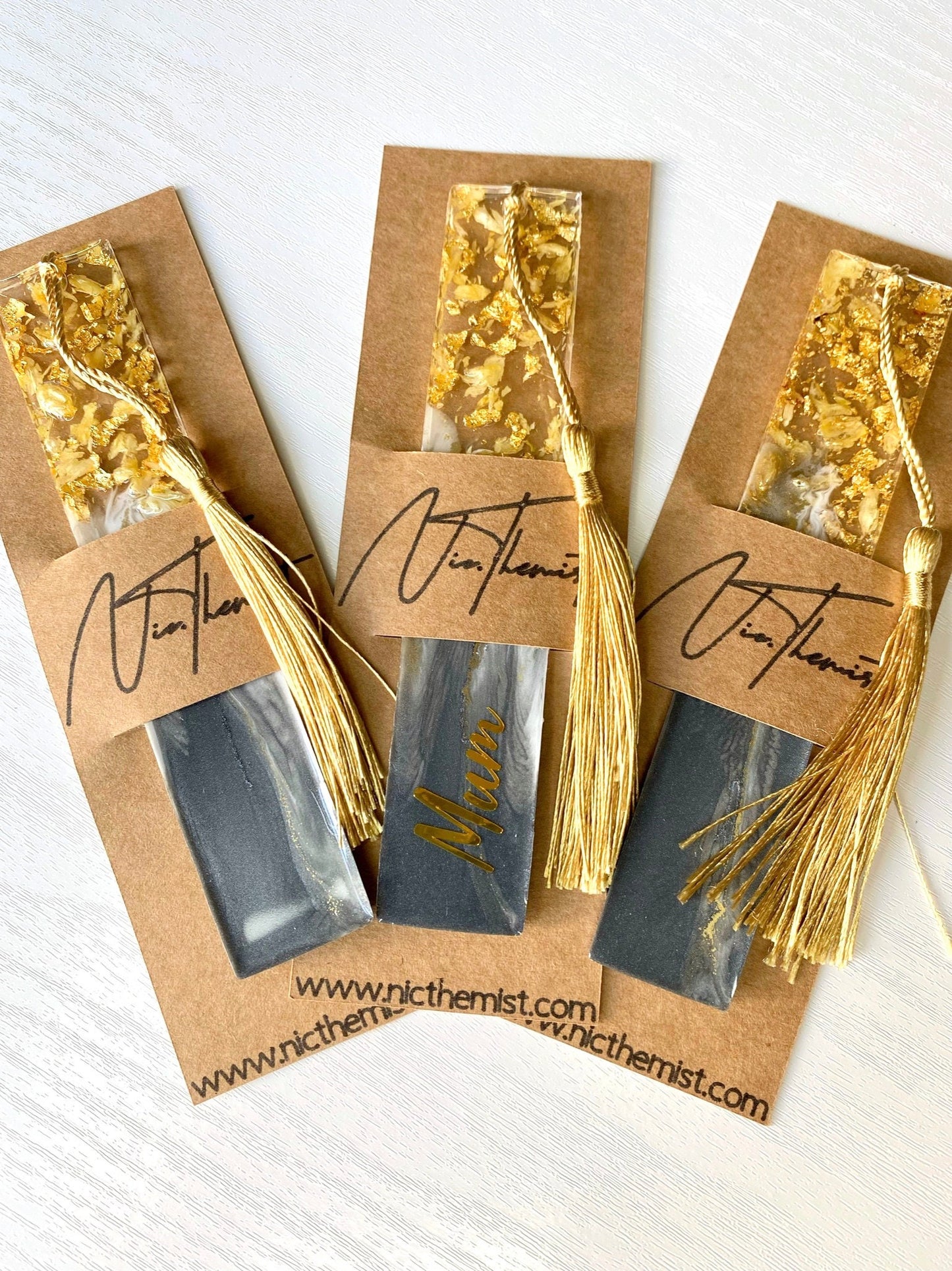 Three grey resin bookmarks with dried jasmine petals and gold leaf inside packaging sleeve with logo, handmade personalized bookmarks for gifts or reading.