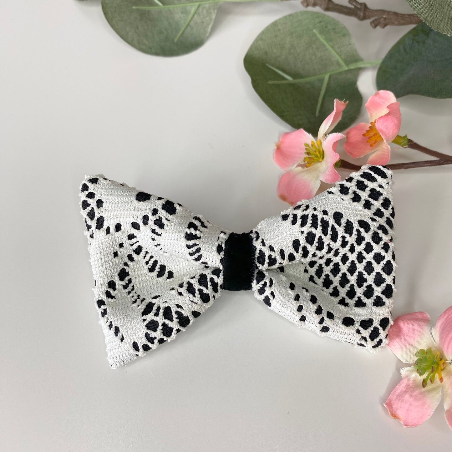 Handmade white and black lace hair bow clips with jacquard jersey fabric and black velvet loop detail, displayed on white surface with flowers in the background