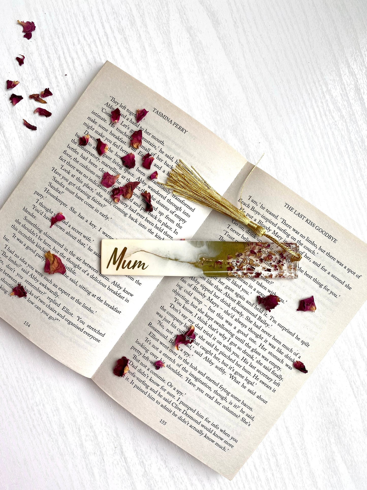 White and green resin bookmark with dried roses and gold leaf, personalised with gold vinyl, placed inside an open book, handmade bookmark for readers or gifts