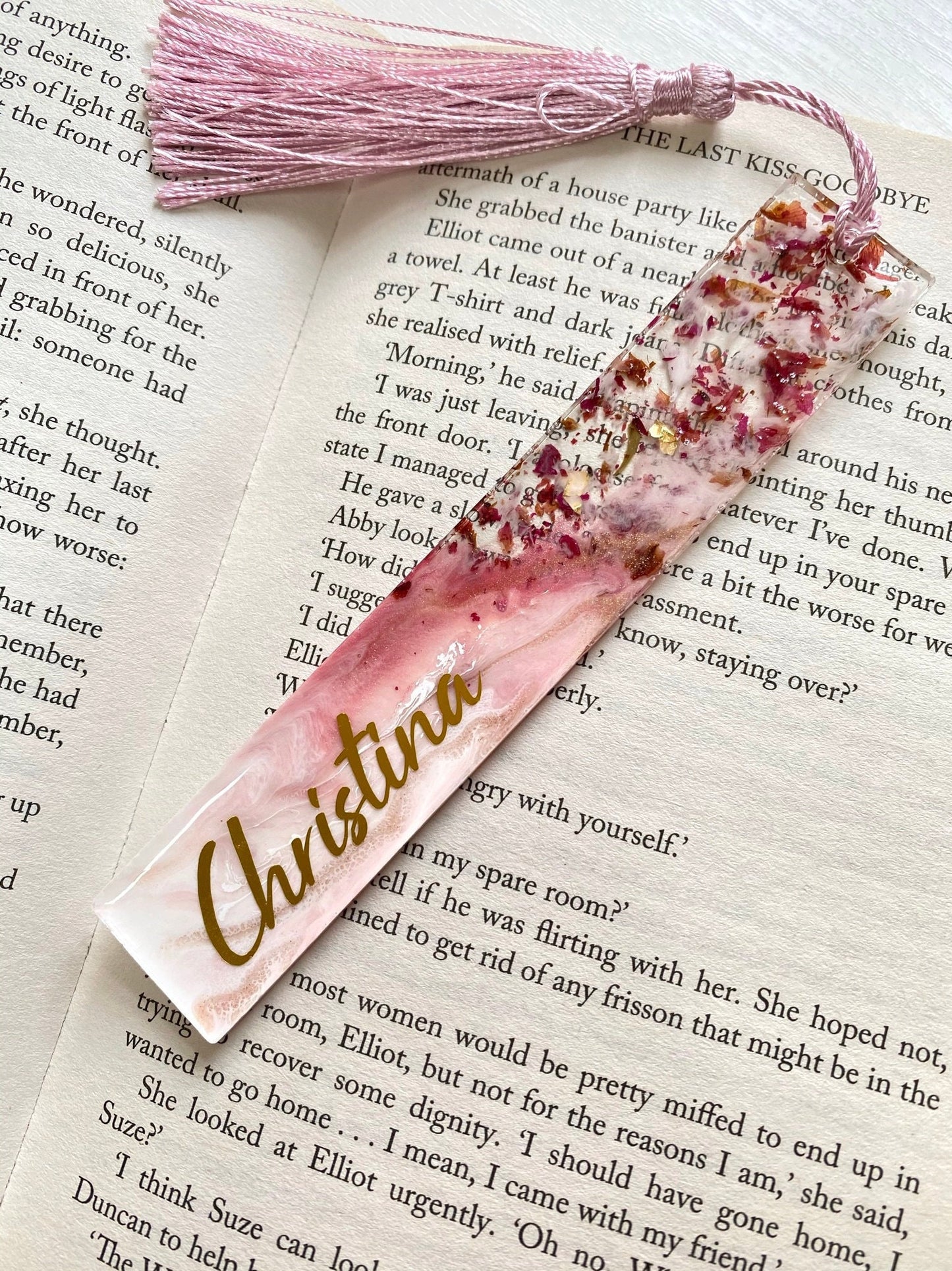 White and pink resin bookmark with dried roses and gold leaf, personalised with gold vinyl, placed inside an open book, handmade bookmark for readers or gifts.