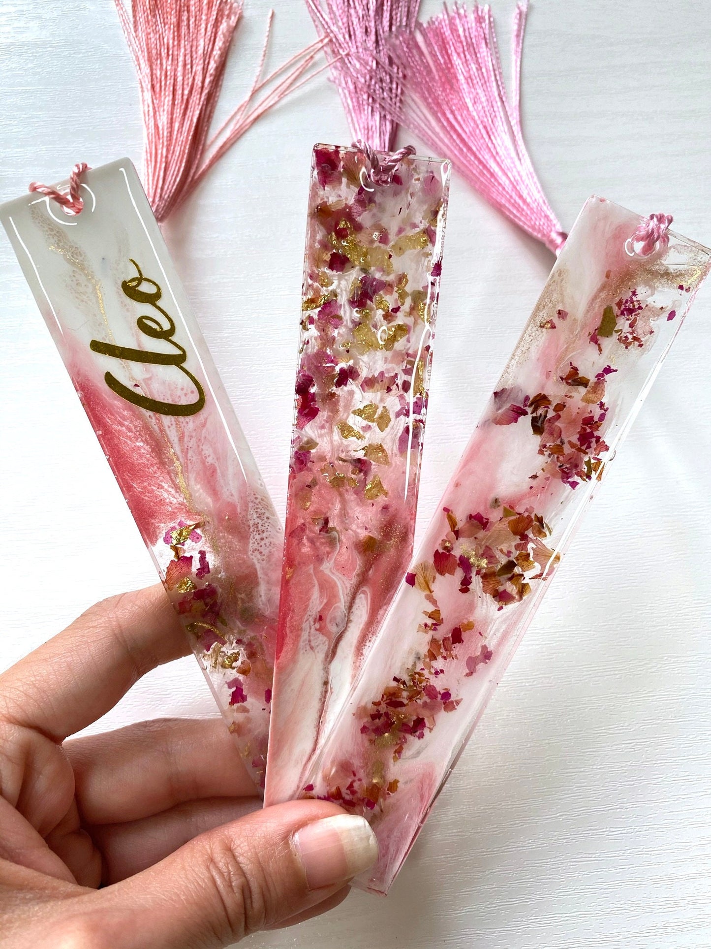 Hand holding three white and pink resin bookmarks with dried roses and gold leaf, including personalised and non-personalised options, handmade bookmarks for readers or gifts