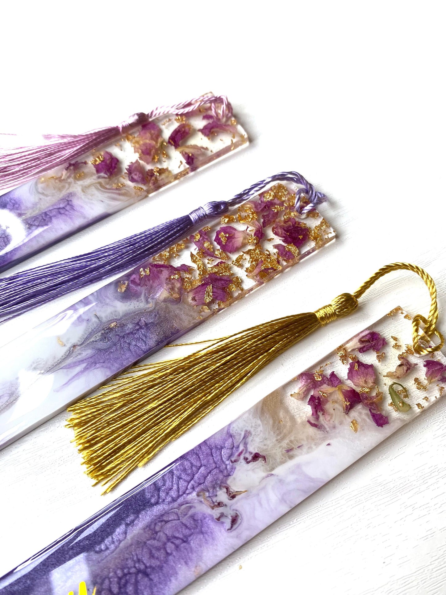 Close-up of three white and purple resin bookmarks with dried roses and gold leaf, one personalised on a purple base, one non-personalised on a white base, and one non-personalised on a purple base, showing handmade details, ideal bookmarks for readers or gifts.