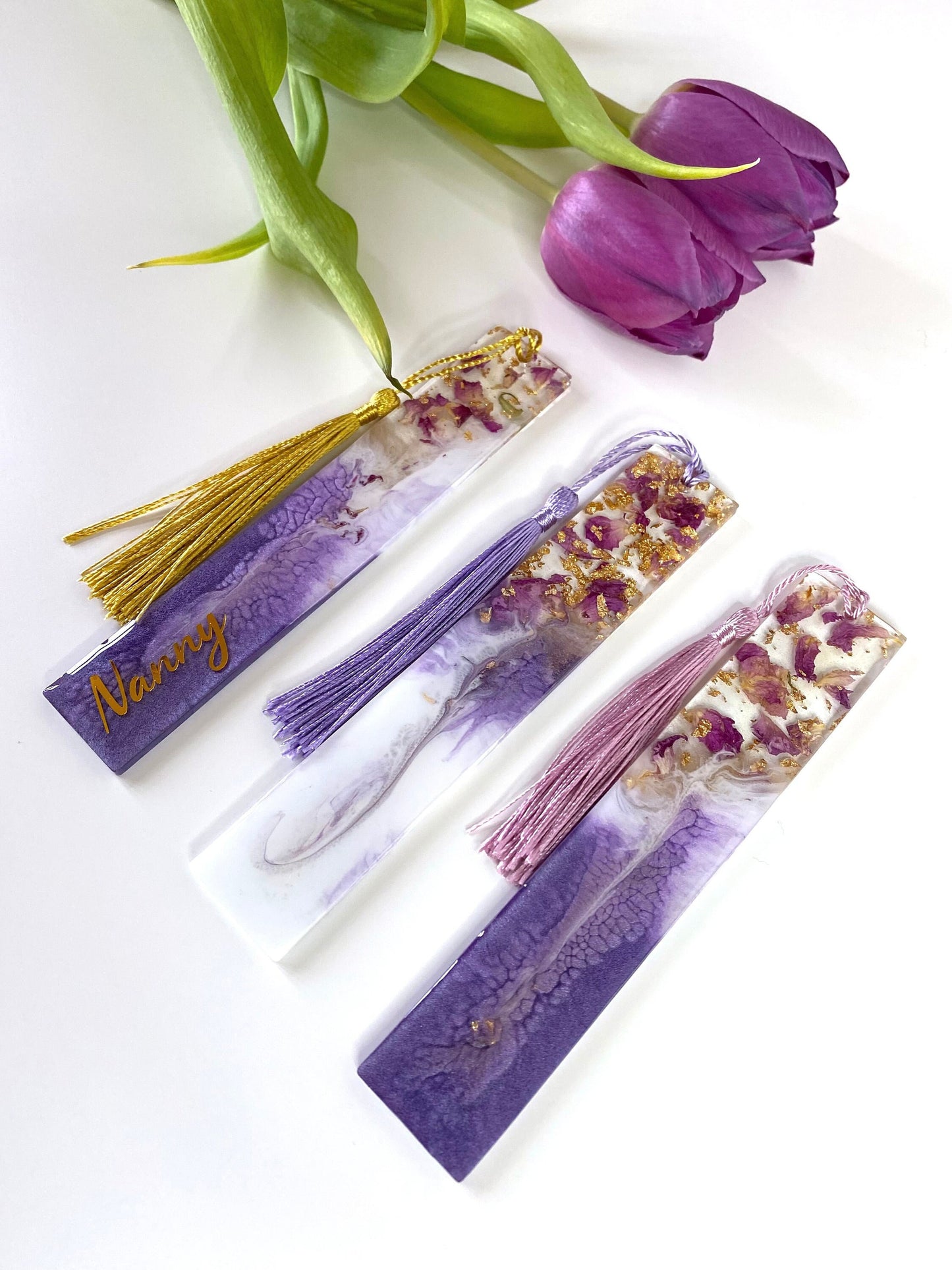 Three white and purple resin bookmarks with dried roses and gold leaf, one personalised with gold vinyl on a purple base, one non-personalised on a white base, and one non-personalised on a purple base, handmade bookmarks for readers or gifts.
