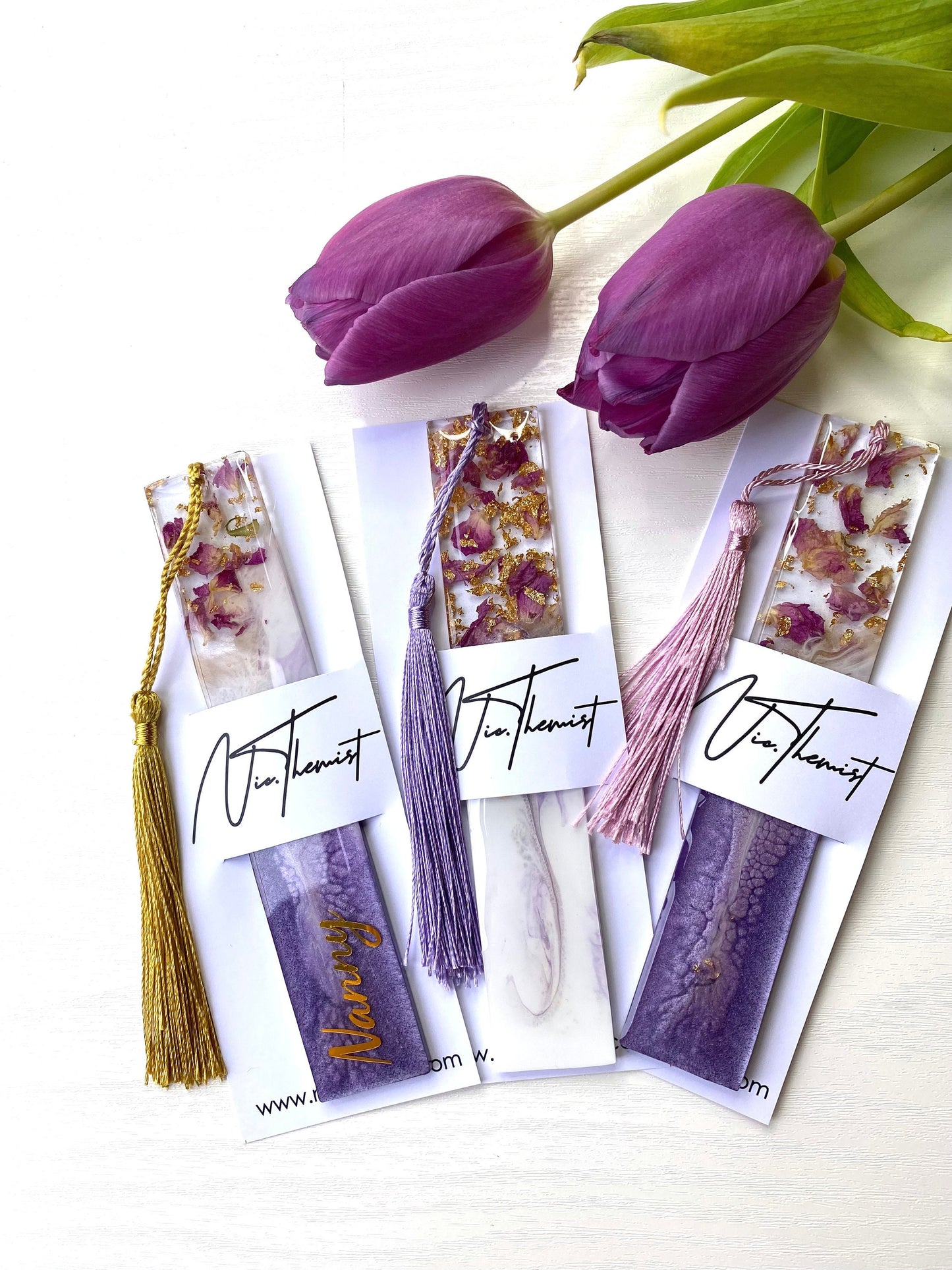 Three white and purple resin bookmarks with dried roses and gold leaf inside packaging sleeves with logo, including one personalised on a purple base, one non-personalised on a white base, and one non-personalised on a purple base, handmade bookmarks for readers or gifts.