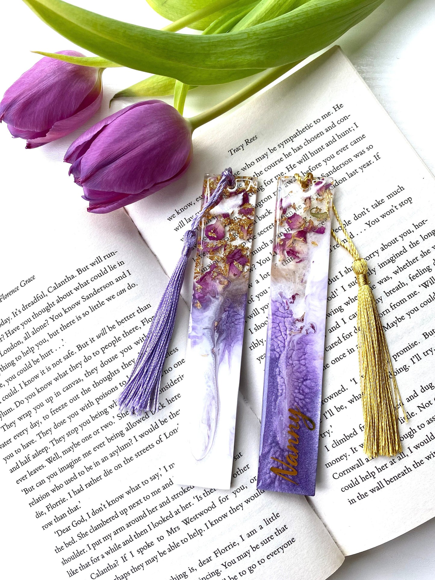 White and purple resin bookmarks with dried roses and gold leaf, both personalised with gold vinyl, placed inside an open book, handmade bookmarks for readers or gifts.