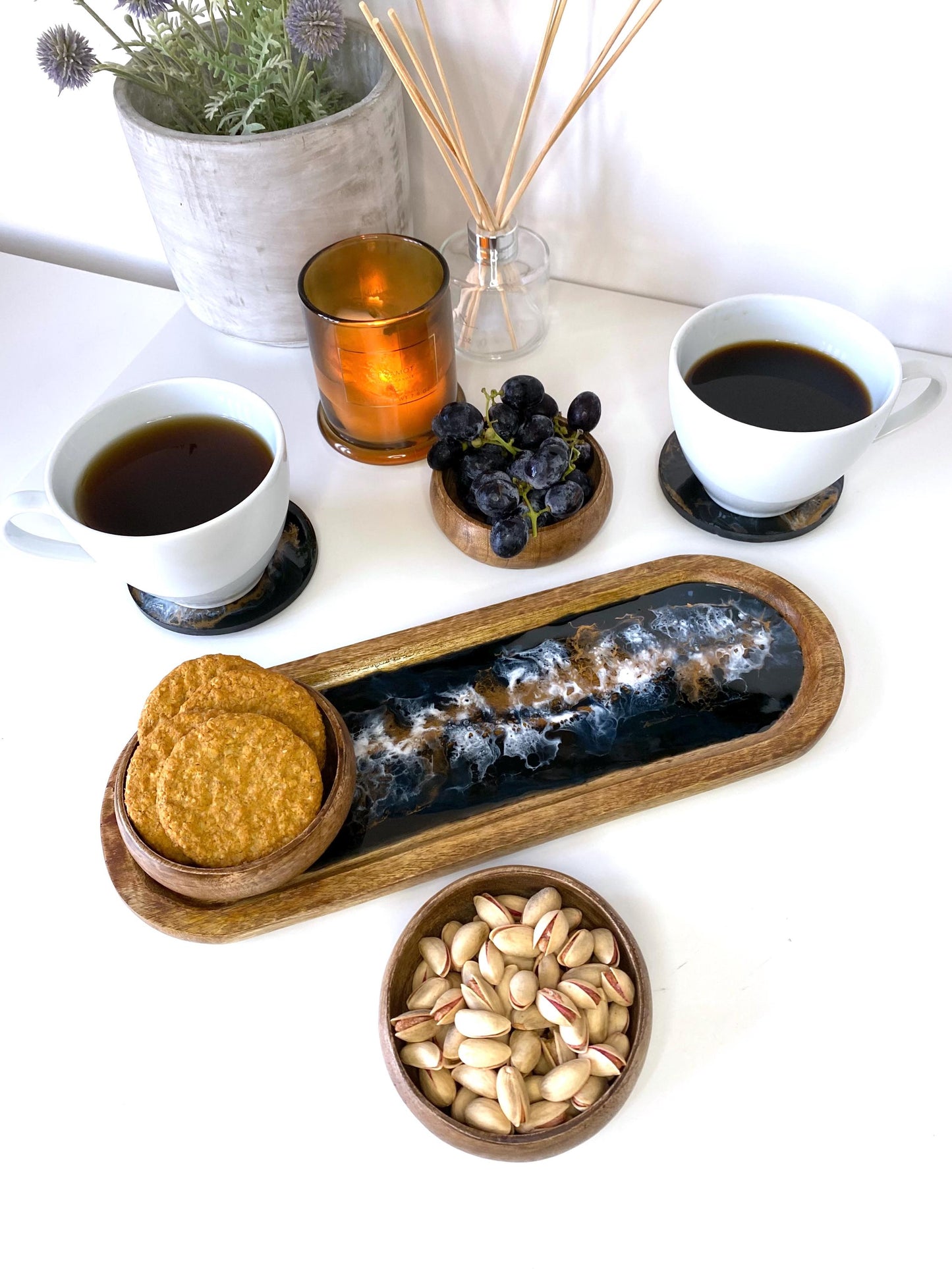 Top-down view of large handcrafted wooden oval tray with epoxy resin details, featuring three wooden bowls with cookies, grapes, and nuts, alongside two matching resin coasters with coffee mugs – functional and stylish serving display & snack platter