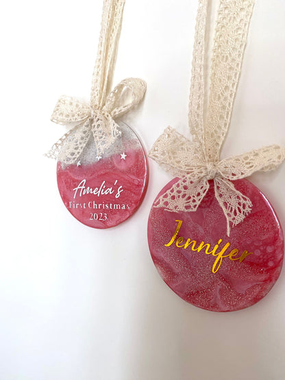 Two Pink Baby First Christmas resin baubles with glittery gold tops and twinkling stars, one personalised in silver lettering with a name, one in gold lettering with "1st Christmas", both hanging with beige lace ribbons and bows