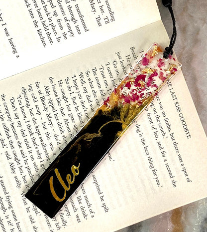 Black and gold resin bookmark personalised with gold vinyl name, placed inside a book, showing dried rose petals in clear resin