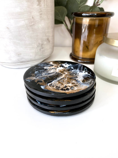 Set of four round resin and slate coasters in black, white and bronze, stacked on top of each other, showing abstract modern design and handmade craftsmanship, decorative coasters for home or gifts.