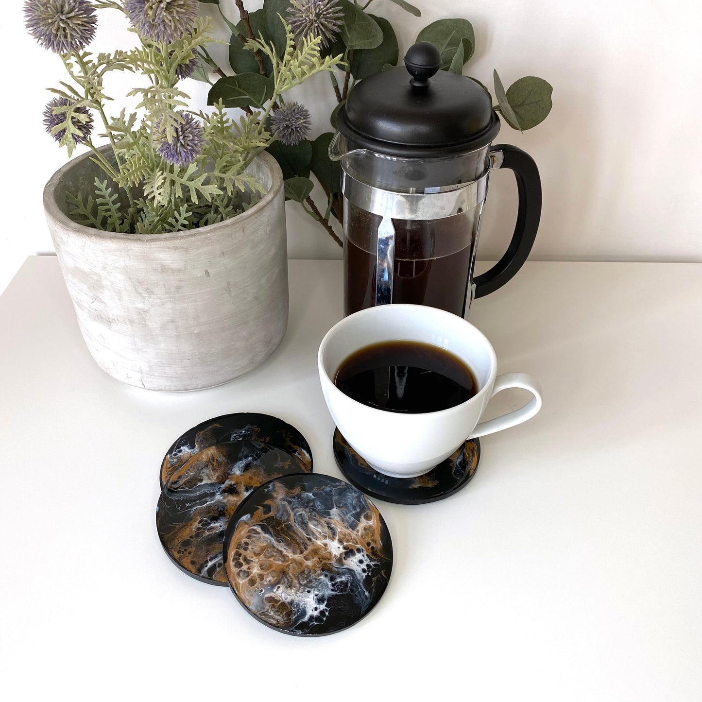 Set of four round resin and slate coasters in black, white and bronze, styled on table with coffee cup on one coaster, filter coffee maker in the background, highlighting abstract modern design, handmade decorative coasters for home or gifts.