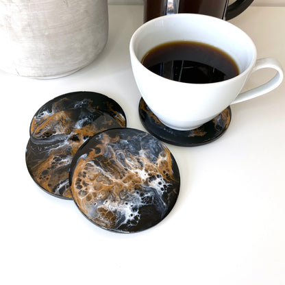 Set of four round resin and slate coasters in black, white and bronze, styled on table with coffee cup on one coaster, three coasters partially stacked with one fully visible to show abstract design, handmade decorative coasters for home or gifts.