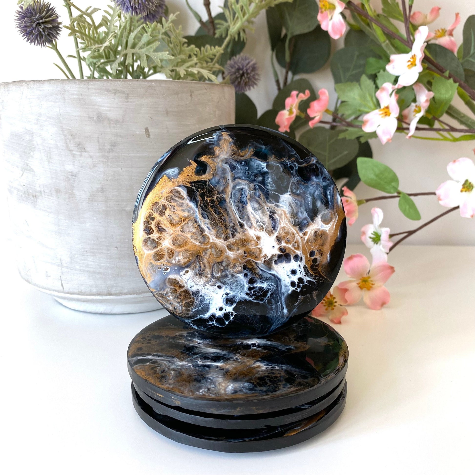 Set of four round resin and slate coasters in black, white and bronze, styled on table with pot and flowers in background, three coasters stacked and one balanced on top to showcase full design, handmade decorative coasters for home or gifts.