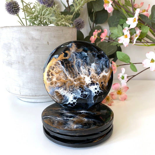 Set of four round resin and slate coasters in black, white and bronze, styled on table with pot and flowers in background, three coasters stacked and one balanced on top to showcase full design, handmade decorative coasters for home or gifts.