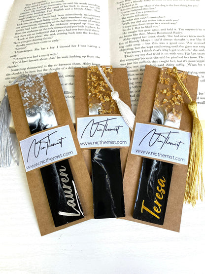 Three black and white resin bookmarks with gold and silver leaf inside packaging sleeves with logo, one personalised in gold vinyl, one personalised in silver vinyl, and one non-personalised, handmade bookmarks for readers or gifts