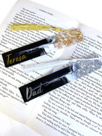 Black and white resin bookmarks with gold and silver leaf, one personalised with gold vinyl and the other with silver vinyl, placed inside an open book, elegant handmade bookmarks for readers or gifts