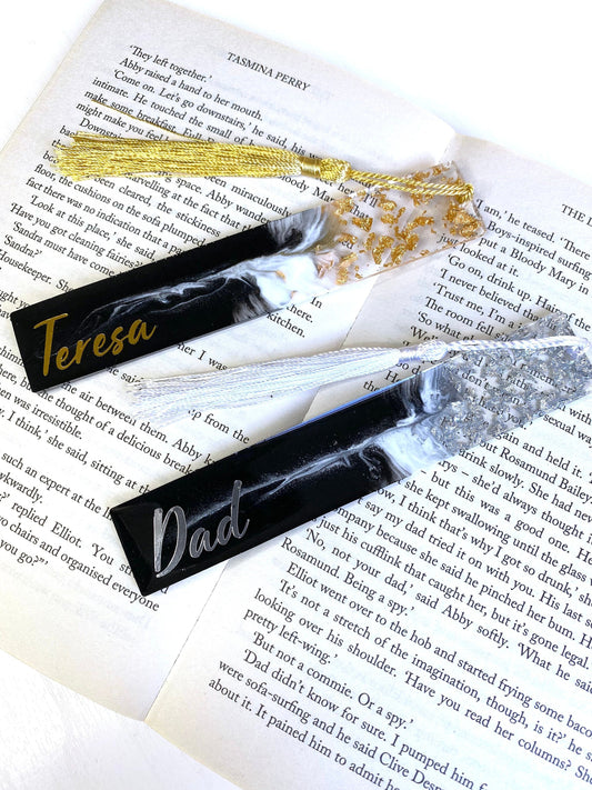 Black and white resin bookmarks with gold and silver leaf, one personalised with gold vinyl and the other with silver vinyl, placed inside an open book, elegant handmade bookmarks for readers or gifts