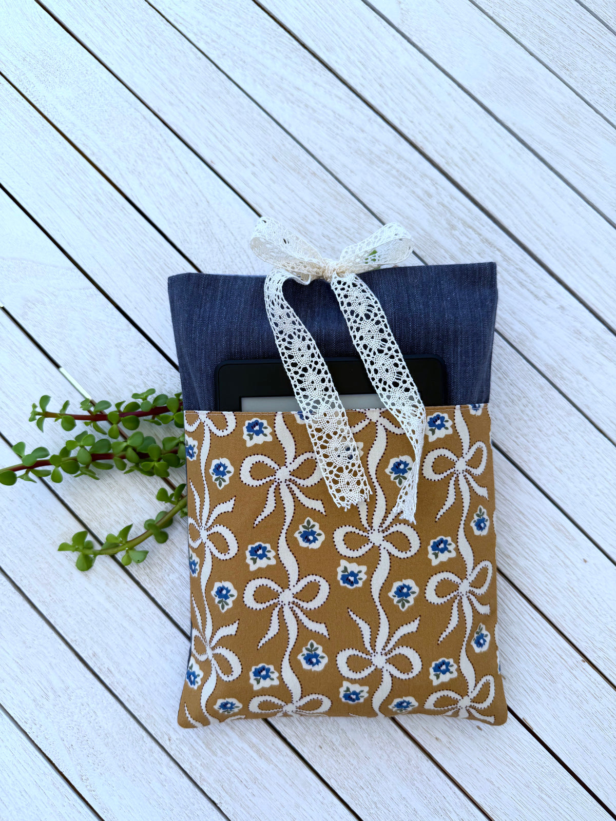 Navy fabric book sleeve with a small black e-reader tucked into its caramel-brown bow print front pocket, showing storage for reading accessories.