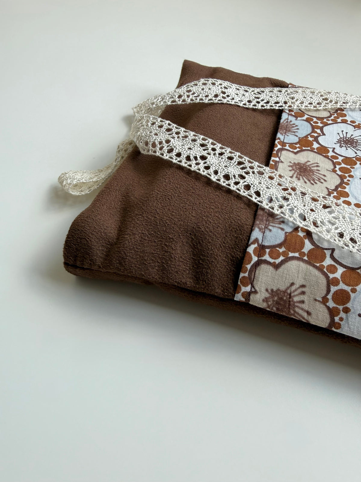 Close-up of the brown faux suede book sleeve folded on a white surface, showing the soft suede-like fabric, retro floral cotton pocket and cream lace ribbon.