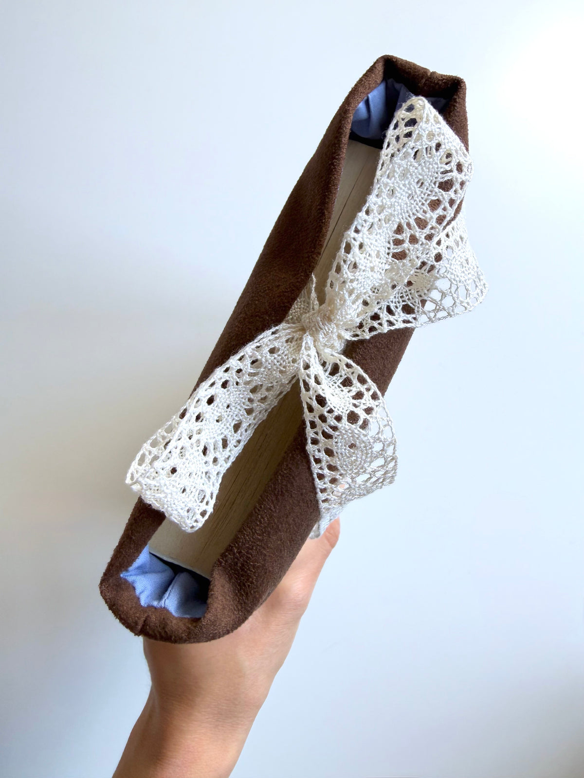 Top view of a handmade brown faux suede book sleeve held in one hand, showing the fabric lining, paperback book inside and cream lace bow closure.