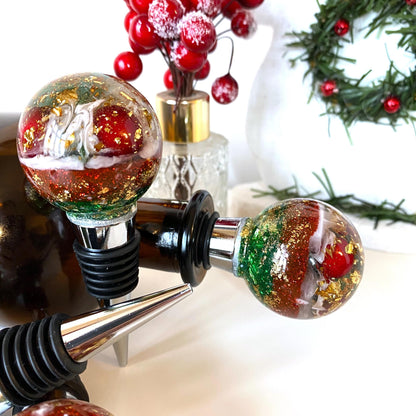 Close-up of Christmas resin wine stopper in gin bottle with two additional stoppers on surface showing red berries, gold leaf, and glitter details