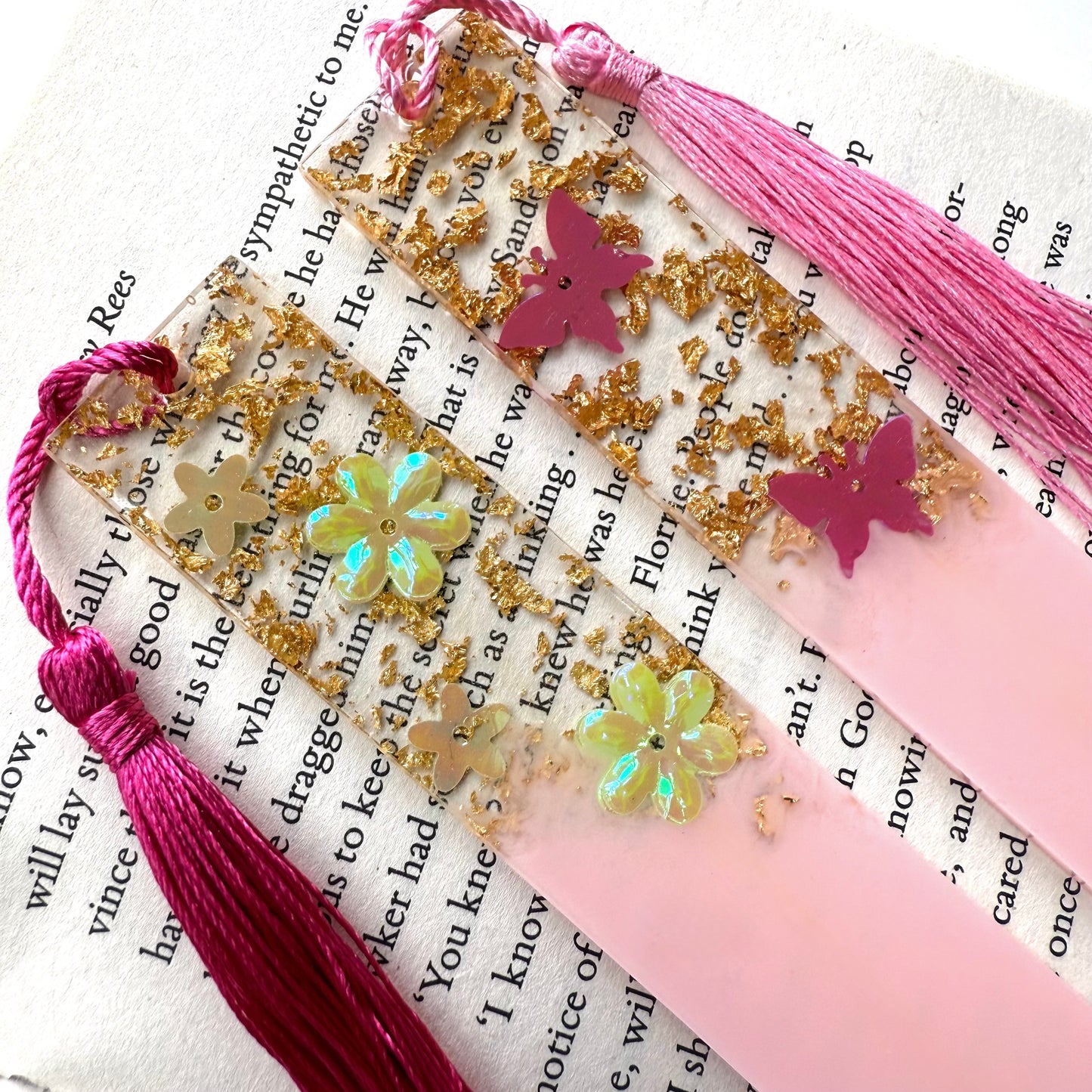 Close-up of two handmade resin bookmarks on an open book: one with butterflies and gold leaf, one with flowers and gold leaf, both with tassels.