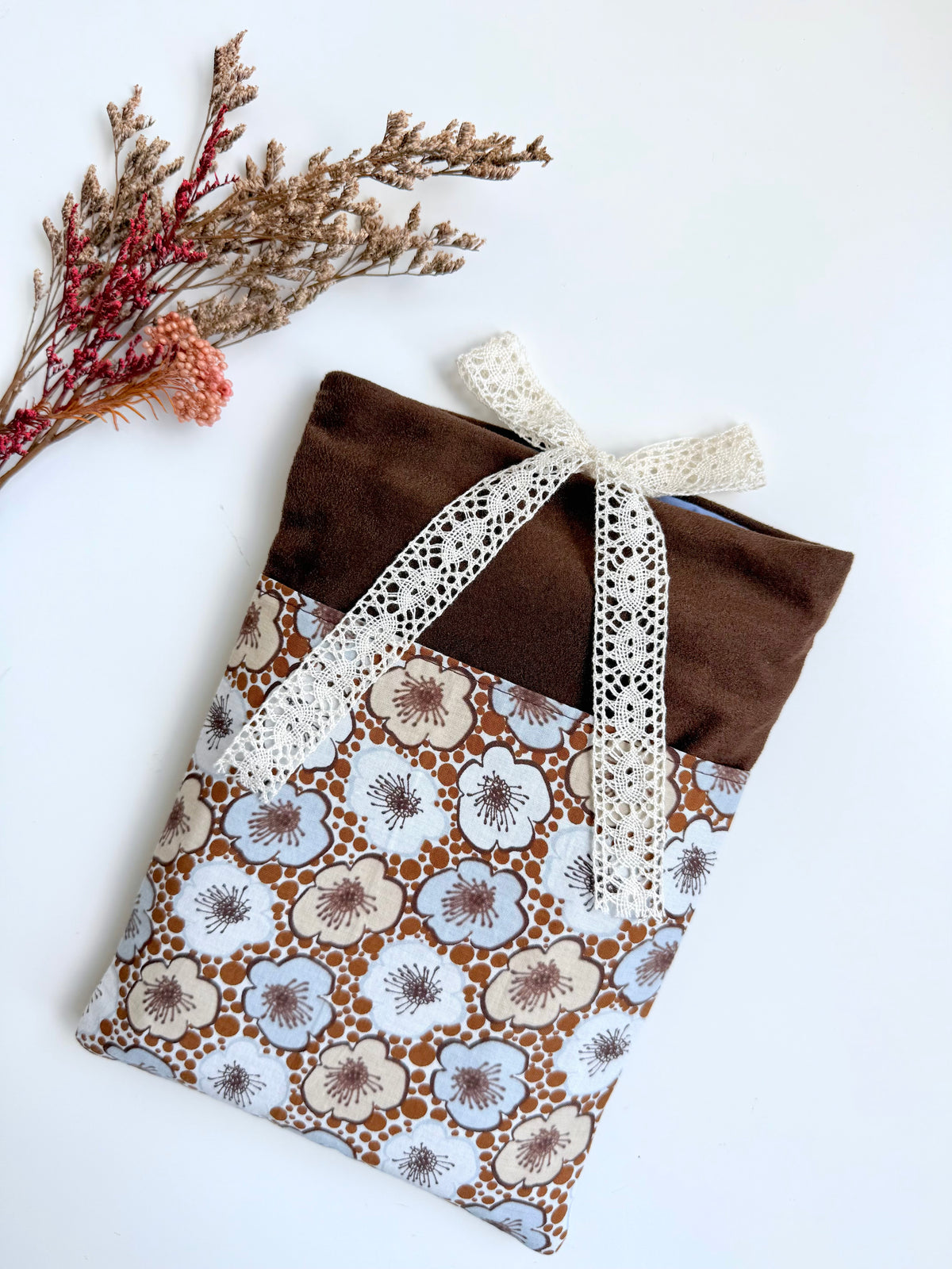 Handmade cottagecore book sleeve featuring chocolate-brown faux suede, a vintage-inspired floral cotton front pocket and cream lace bow, photographed with dried flowers.