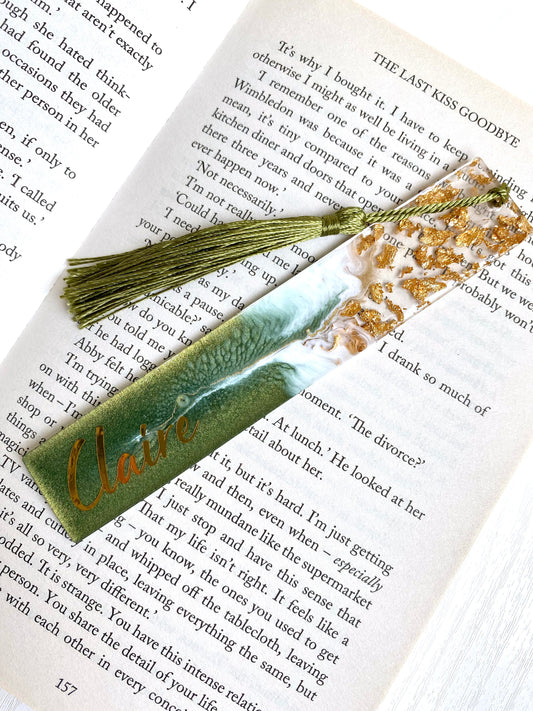 Green resin bookmark with gold leaf, personalised with gold vinyl, placed inside an open book, handmade bookmark for readers or gifts