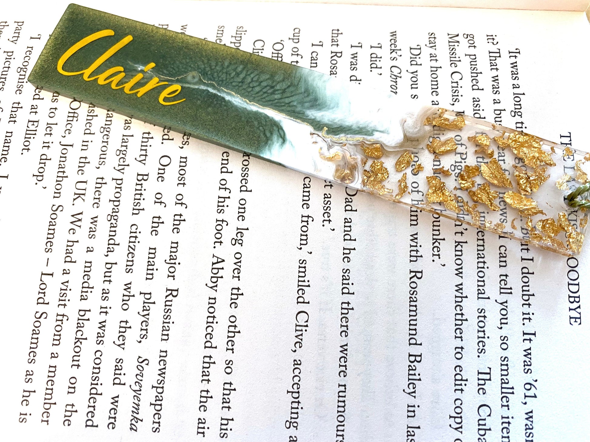 Green resin bookmark with gold leaf, personalised with gold vinyl, placed inside an open book, handmade bookmark for readers or gifts.