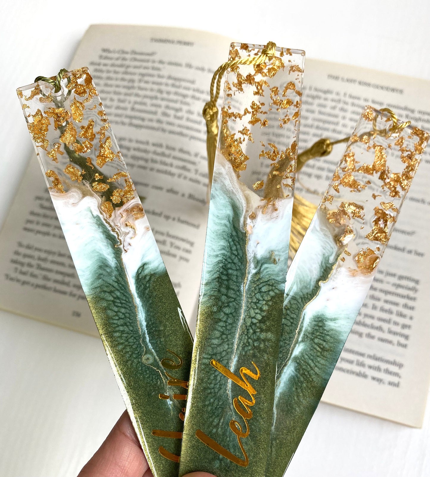 Hand holding three green resin bookmarks with gold leaf, including personalised and non-personalised options with colourful tassels, handmade bookmarks for readers or gifts