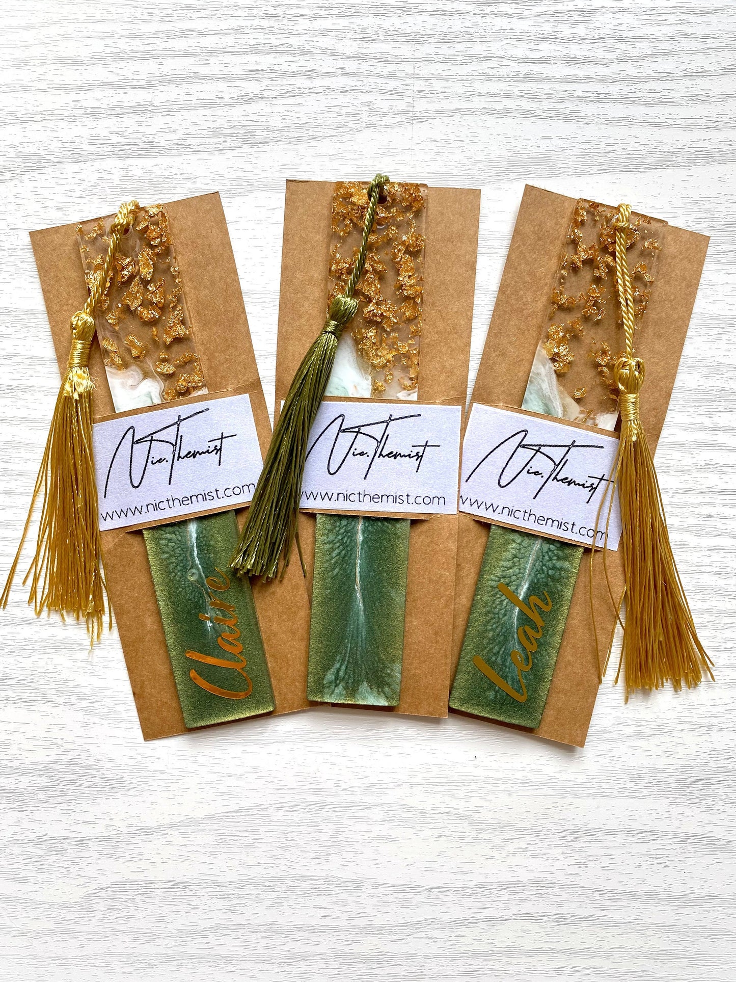 Three green resin bookmarks with gold leaf inside packaging sleeves with logo, including personalised and non-personalised options, each with colourful tassels, handmade bookmarks for readers or gifts.