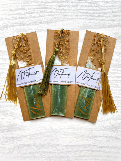 Three green resin bookmarks with gold leaf inside packaging sleeves with logo, including personalised and non-personalised options, each with colourful tassels, handmade bookmarks for readers or gifts.