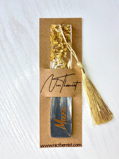 Grey resin bookmark with dried jasmine petals and gold leaf, personalised with gold vinyl, inside packaging sleeve with logo, featuring a gold tassel, handmade gift bookmark for readers