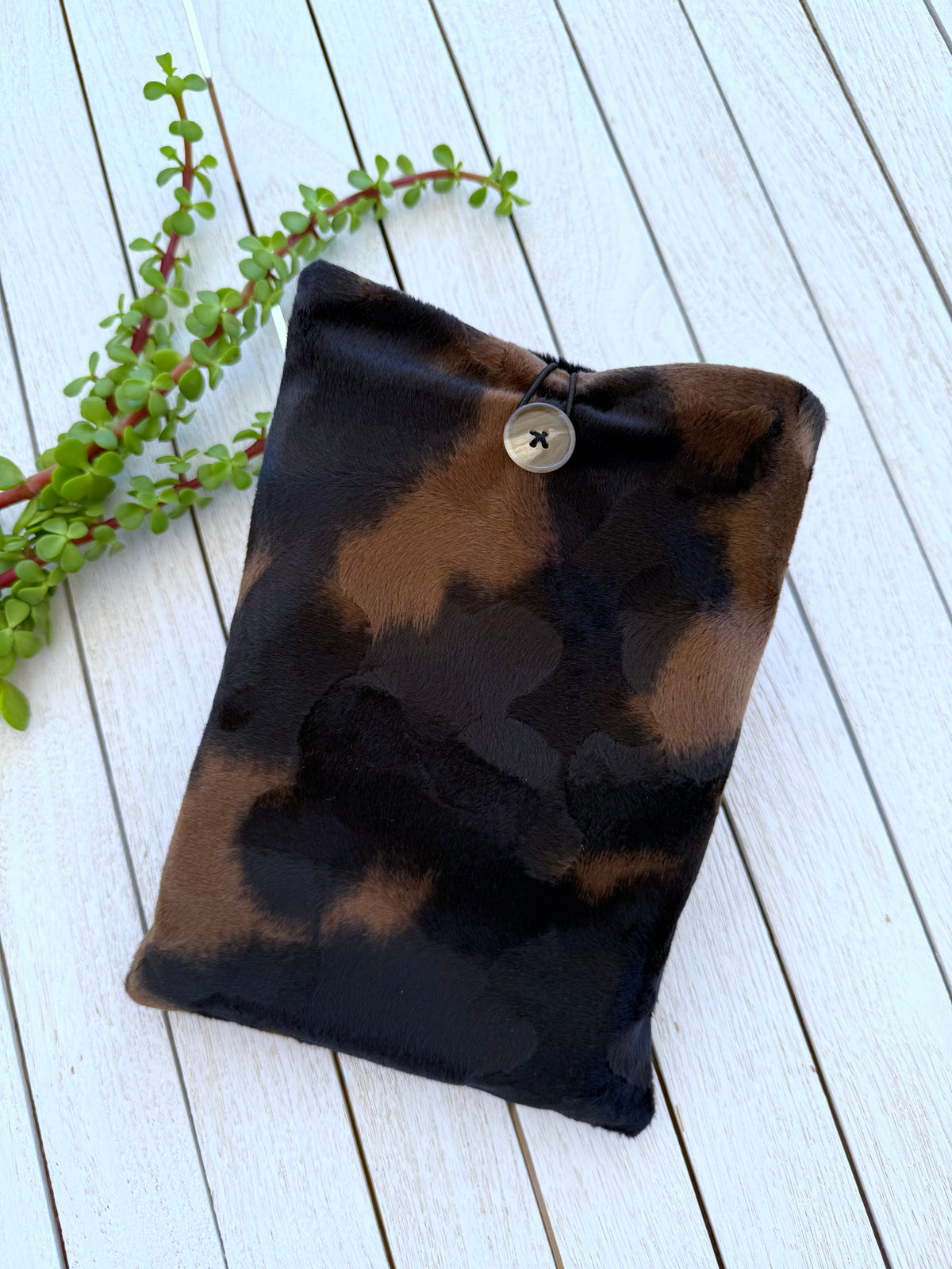 Full front view of a black and brown animal print velvet book sleeve with button closure, photographed on white wooden boards with greenery.