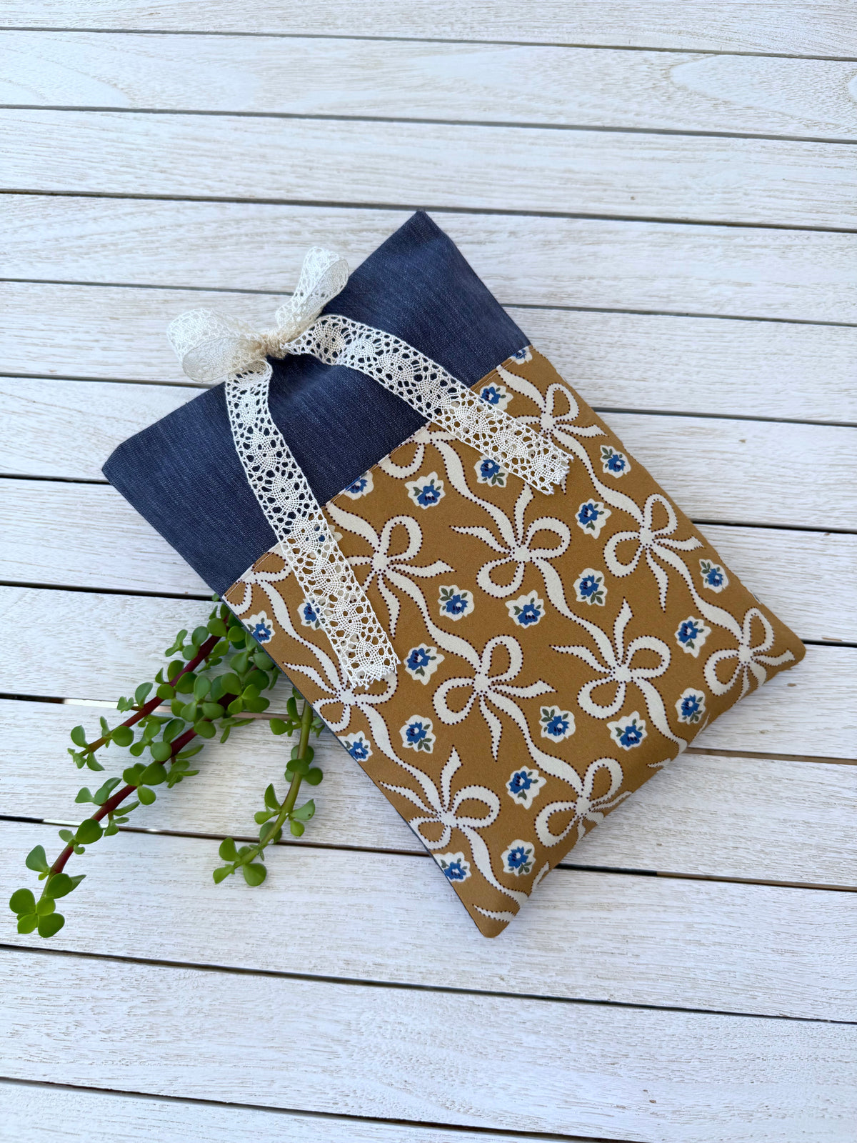 Handmade navy book sleeve with a caramel-brown bow print front pocket and cream lace ribbon, displayed diagonally on white wooden boards.