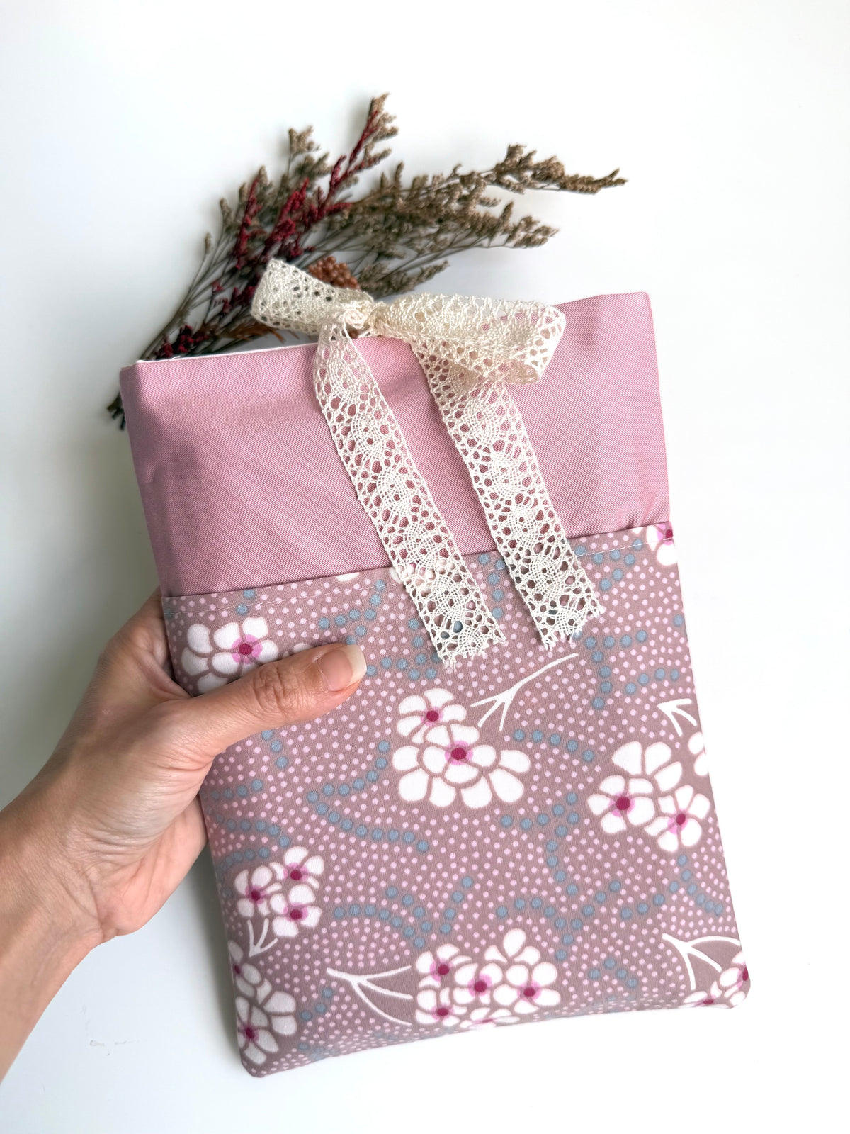 Hand holding a handmade dusty pink fabric book sleeve with a contrasting floral front pocket and cream lace ribbon bow, showing its size and design.