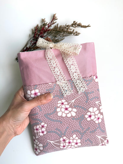 Hand holding a handmade dusty pink fabric book sleeve with a contrasting floral front pocket and cream lace ribbon bow, showing its size and design.