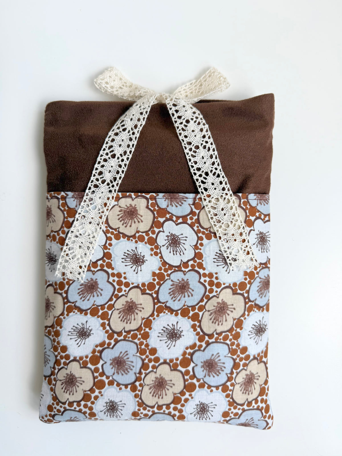 Front view of a handmade brown faux suede book sleeve with cream and pale blue retro floral cotton pocket and cream lace bow closure.