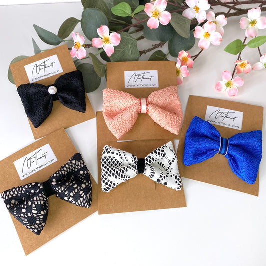 Handmade lace hair bow clips in black, blue, white, and peach attached to branded packaging sleeves, displayed on white surface with flowers in background