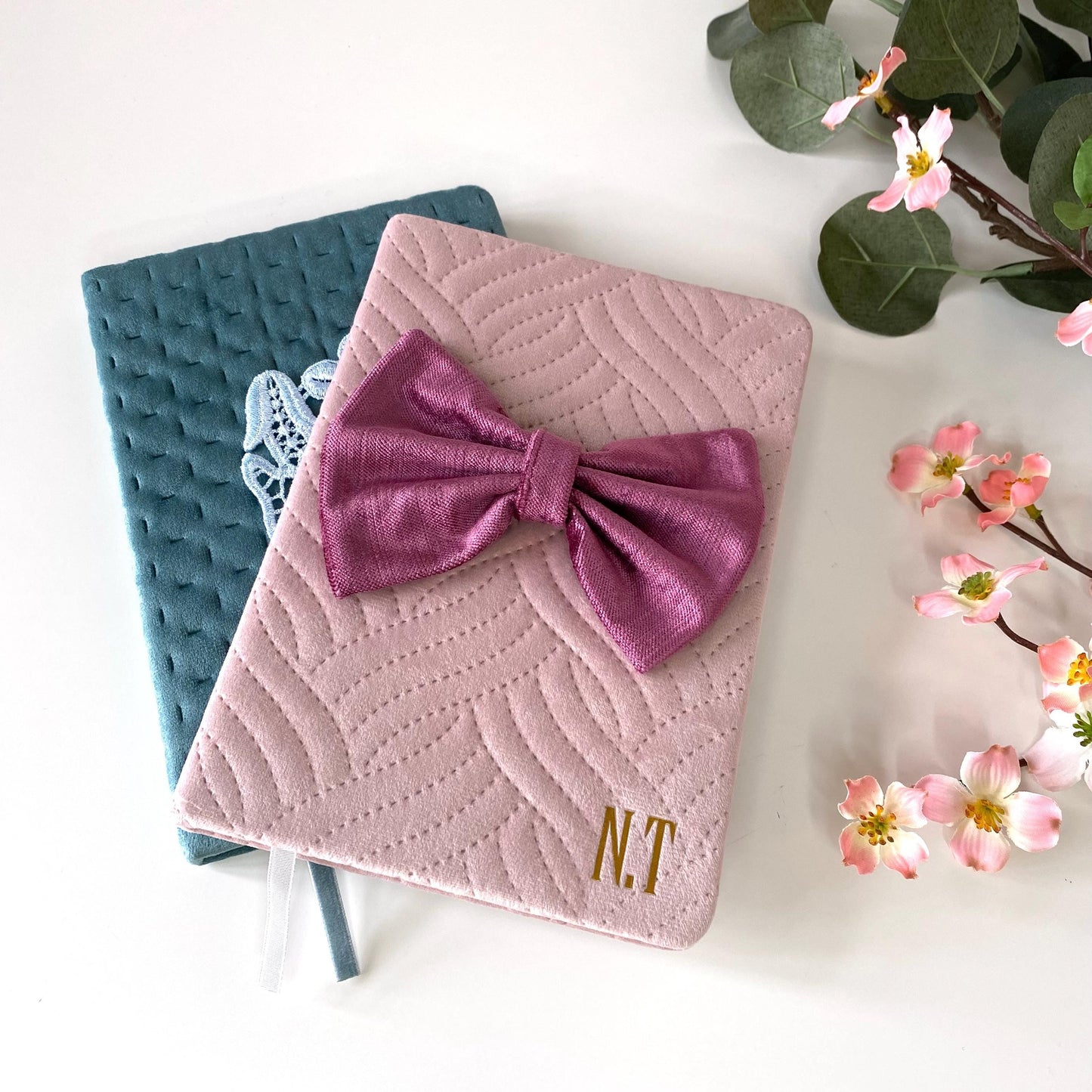 Pink A5 luxury velvet notebook with quilted cover and hand-sewn bow, personalised with gold initials, partially covering teal notebook with floral appliqué, displayed on a white surface with flowers