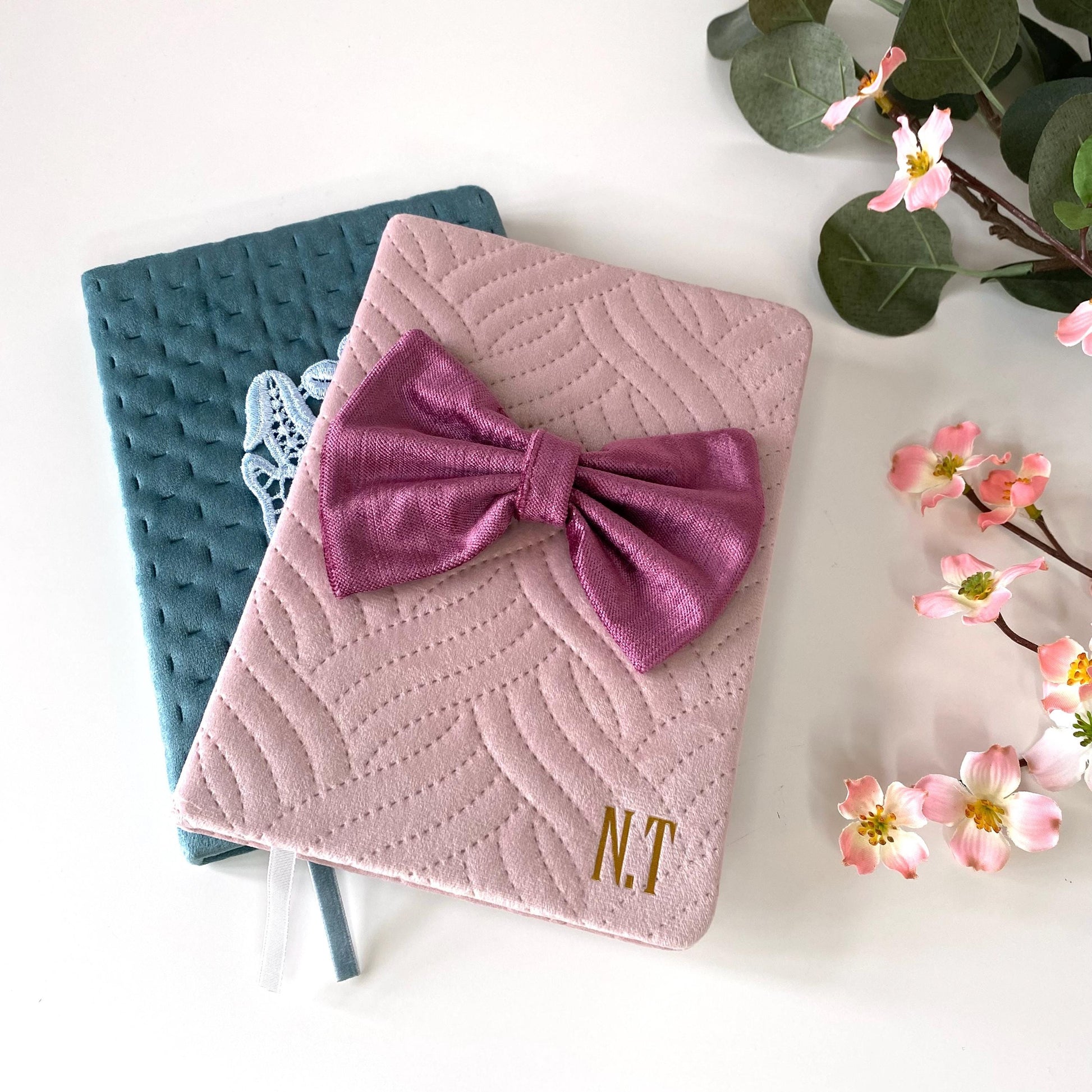 Pink A5 luxury velvet notebook with quilted cover and hand-sewn bow, personalised with gold initials, partially covering teal notebook with floral appliqué, displayed on a white surface with flowers