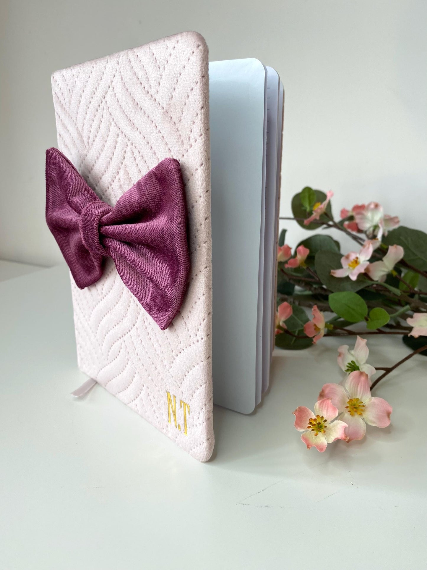 Pink A5 luxury velvet notebook with quilted cover and hand-sewn bow, half open on a white surface showing lined pages, personalisable with initials