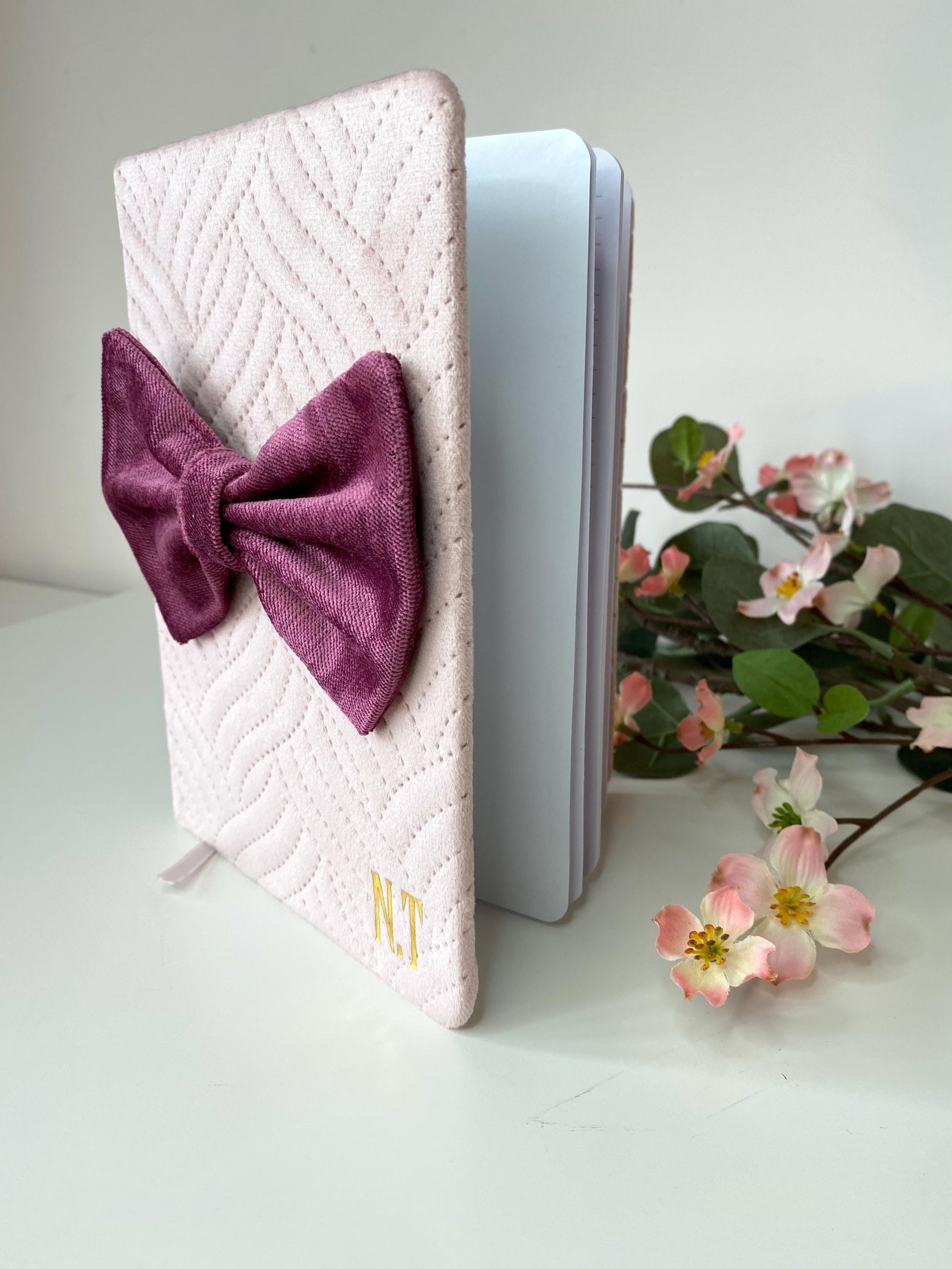 Pink A5 luxury velvet notebook with quilted cover and hand-sewn bow, half open on a white surface showing lined pages, personalisable with initials