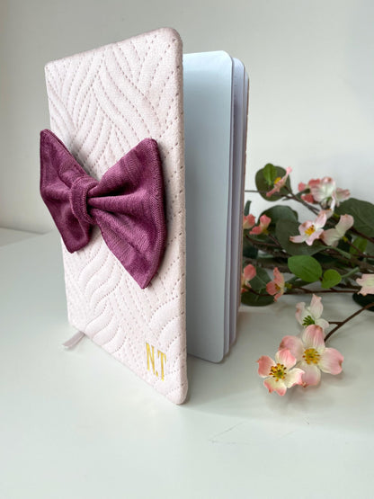 Pink A5 luxury velvet notebook with quilted cover and hand-sewn bow, half open on a white surface showing lined pages, personalisable with initials