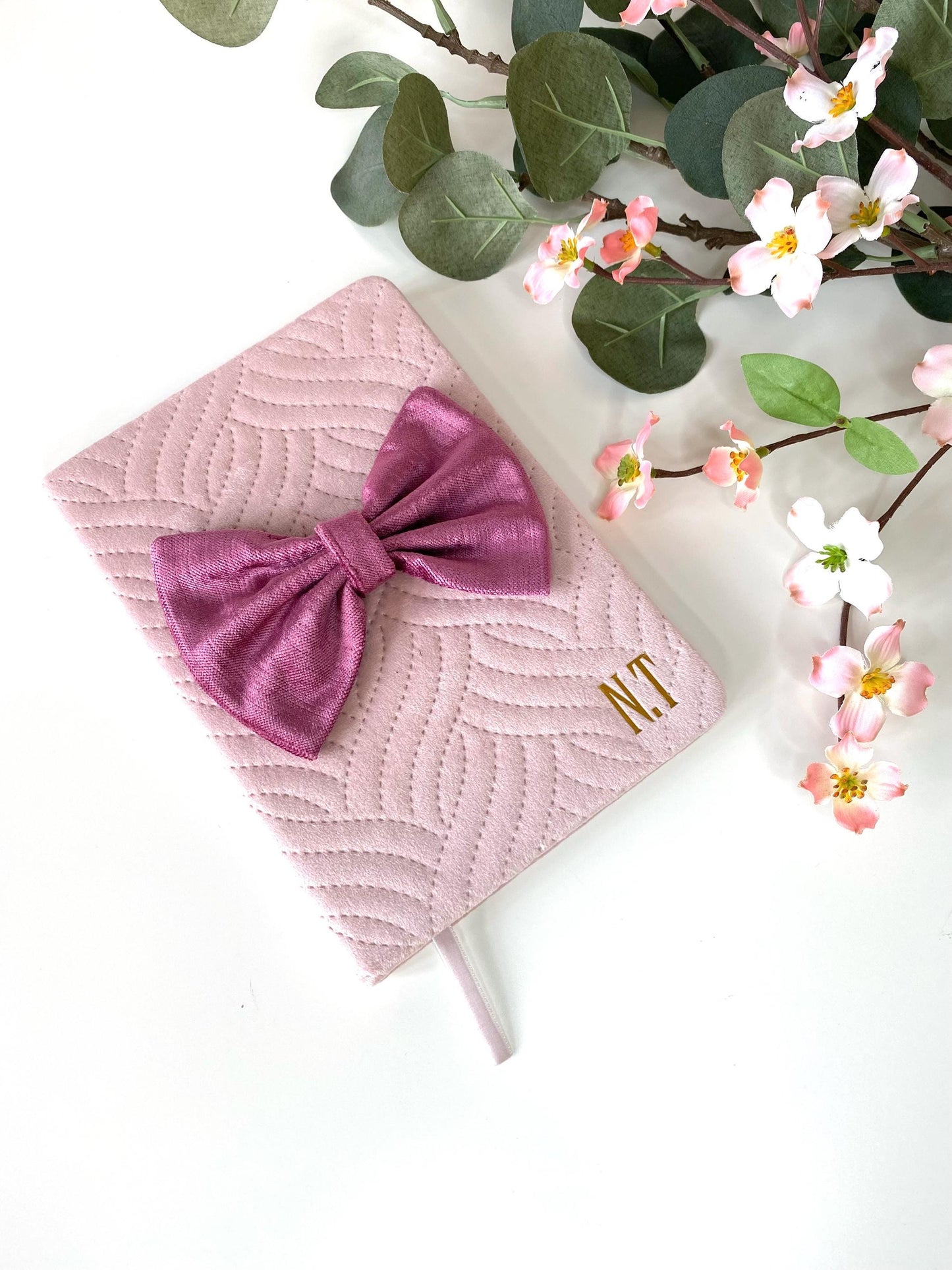 Top-down view of pink A5 luxury velvet notebook with quilted cover and hand-sewn bow, personalised with gold initials, flowers in the background on a white surface