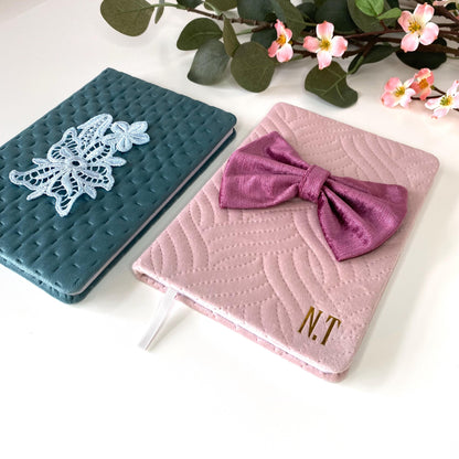 Angled view of A5 luxury velvet notebooks on a surface; teal notebook with floral appliqué and pink notebook with hand-sewn bow, both quilted and personalised