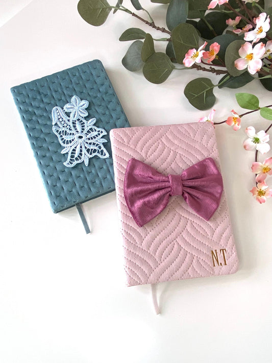A5 luxury velvet notebooks on a white surface with flowers; teal notebook with floral appliqué and pink notebook with hand-sewn bow, both personalisable
