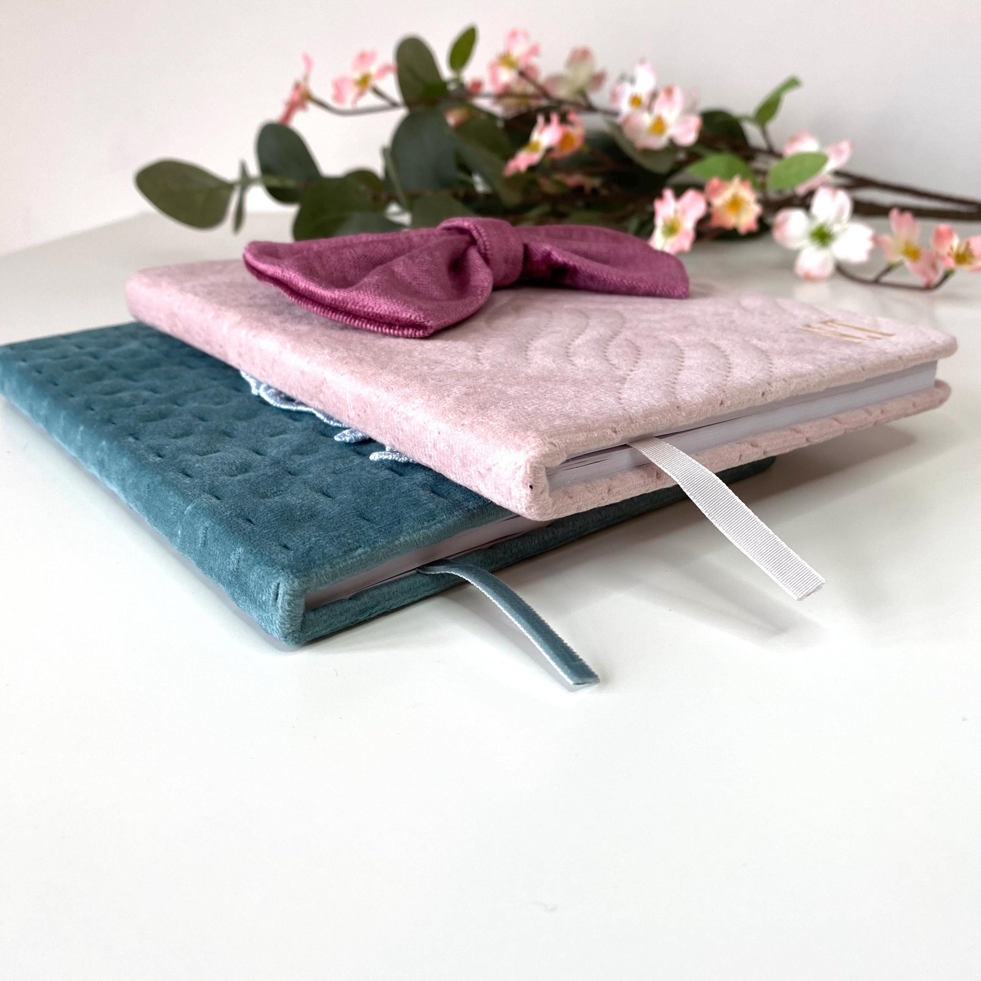 Bottom-angle view of A5 luxury velvet notebooks on a white surface with flowers, showing thickness and quilted covers; pink notebook with bow and teal notebook with floral appliqué, both personalised