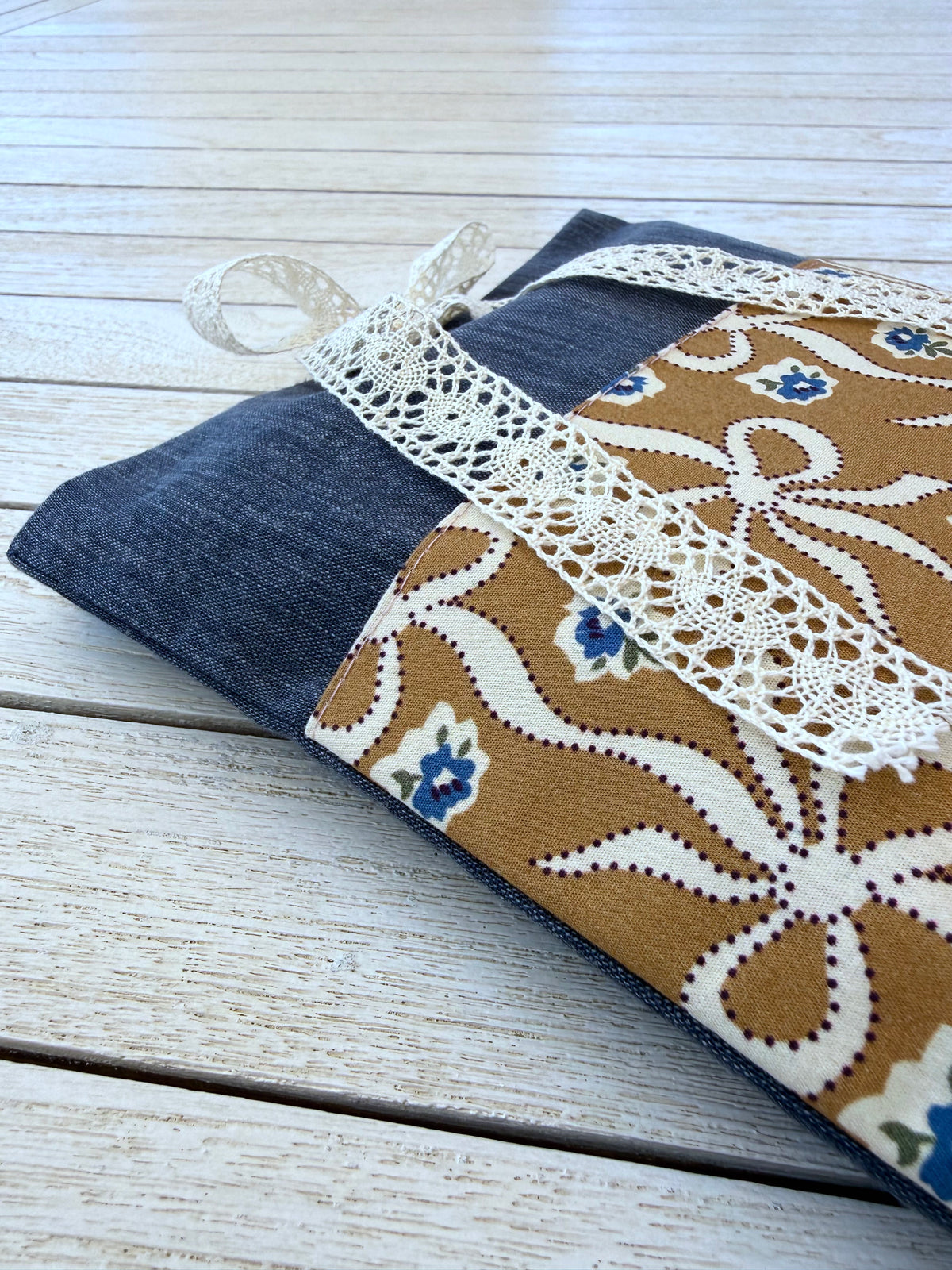 Close-up of a handmade navy denim-look book sleeve lying on white wooden boards, showing the caramel-brown pocket with cream bow patterns, small blue flowers and cream lace ribbon.
