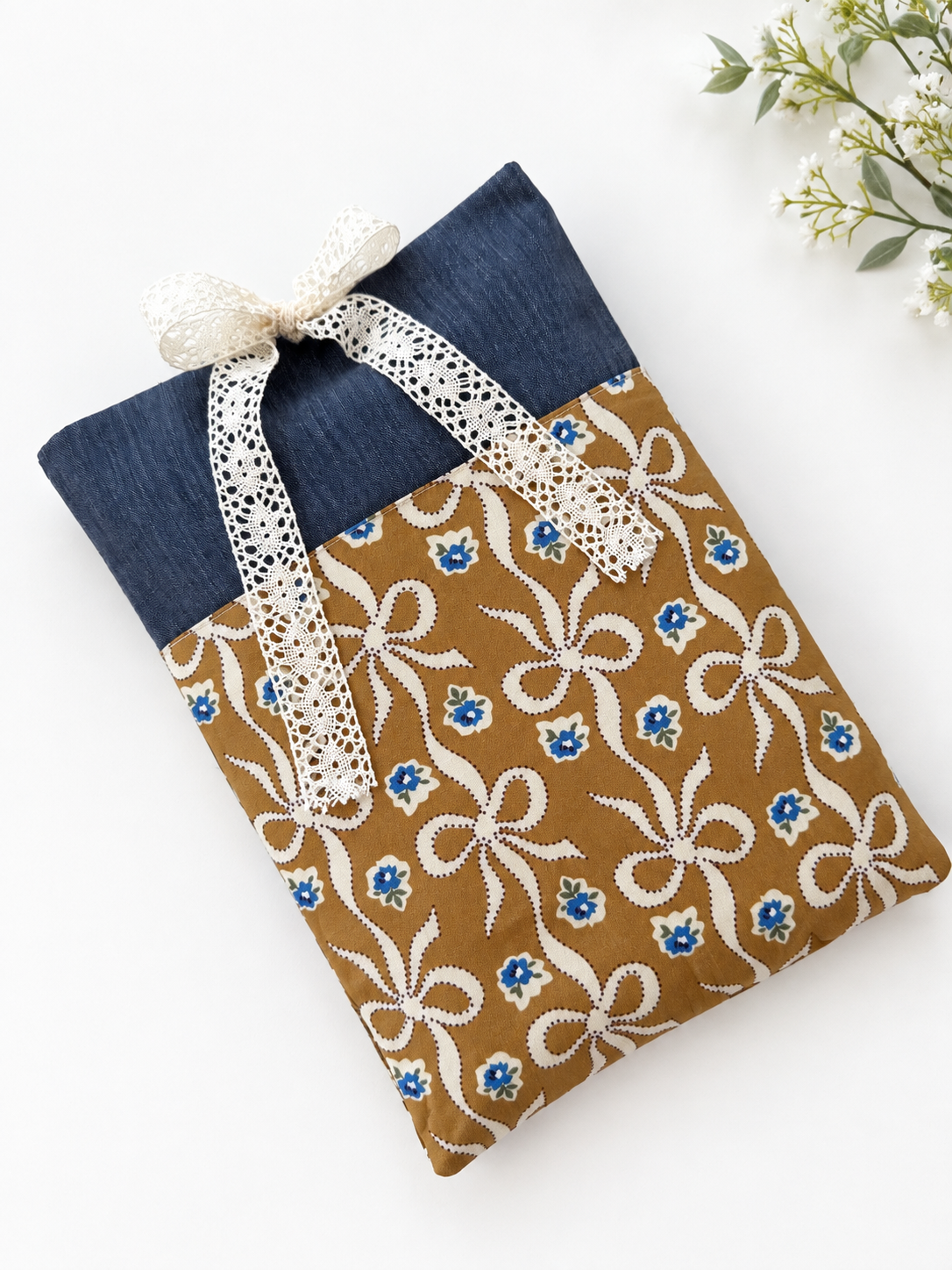 Handmade navy denim-look book sleeve with a caramel-brown front pocket featuring cream bows and small blue flowers, finished with a cream lace ribbon closure.