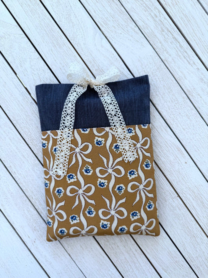 Front view of a handmade navy and caramel-brown book sleeve laid on white wooden boards, showing the cream lace bow closure and decorative bow print pocket.